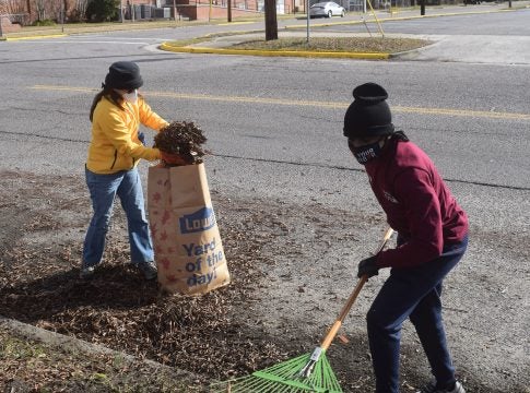 Two children pictured picking up leaves and other debris. One child wields a green rake; the other child is dumping a pile of debris into a large paper bag of outdoor waste.