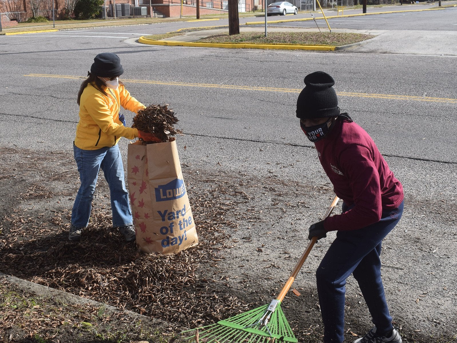 Two children pictured picking up leaves and other debris. One child wields a green rake; the other child is dumping a pile of debris into a large paper bag of outdoor waste.